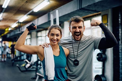 Happy athletic woman and her fitness instructor flexing muscles in a gym while looking at camera.