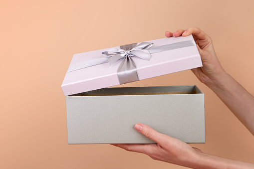 Side view of female hands opening gray purple box with satin ribbon and bow. Gift is hidden inside on a peach beige background. Open or close surprise color trend 2024