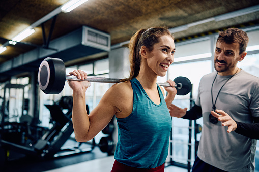Happy athletic woman exercising with barbell with coach's assistance in a gym.