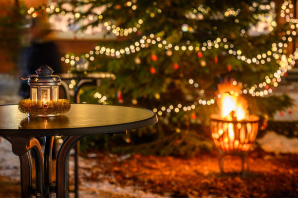 Lantern stands on the table next to a decorated Christmas tree, while a comforting fire flickers in the background. Lantern with festive touch illuminated with festive lights and warm glow of fire.