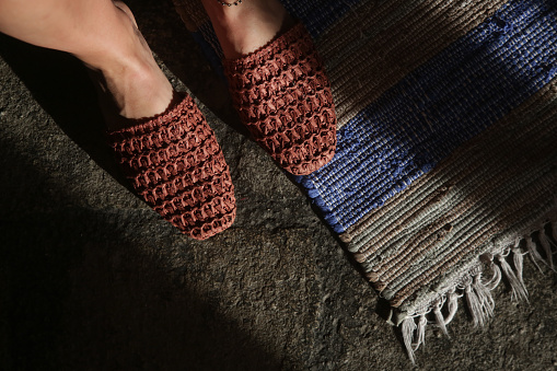 Woman standing in handmade raffia mules on the striped handwoven wool rug. Sustainable ethically made pair of slippers. Responsibly sourced shoes with eco-friendly materials.