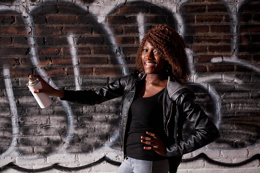 Young black woman (20s) holding paint can, standing in front of brick wall covered with grafitti.