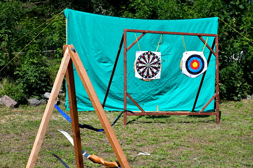 A close up on a set of archery practice targets located against a green protective cloth and with a frame on which bows and crossbows are located together with some arrows seen on a sunny summer day
