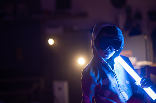 Portrait of boy with hood and VR glasses holding LED lamps at home at night