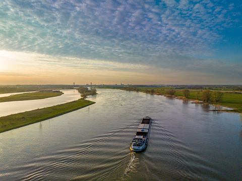 Panoramic aerial view on a freight ship sailing on the river IJssel during a springtime sunset in Overijssel. The flow of the river is leading towards the setting sun in the distance while lights are popping up in the city at the end of a beautiful springtime day.