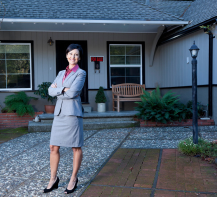 smiling real estate agent standing in front of a home with a FOR SALE sign