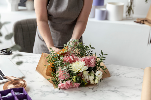 Portrait of female florist in apron arranging fresh flowers for bouquet in the flower shop, using roses, hydrangea, peonies, then wrapping it into color paper and finishing with ribbon