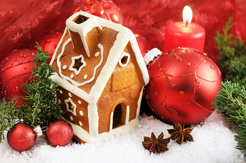 Small gingerbread house on white snow with pine twig and Christmas ornament. In background burning candles and some more Christmas balls. Useful as Christmas and Advent greeting card background.