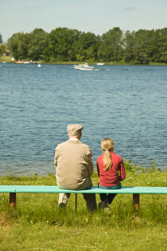 Back view of a grey-haired man holding a curly woman over the shoulders while sitting on a wooden bench next to a calm pond. Copy space. Only heads and shoulders visible.