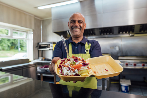 Man running a small takeaway business in North Yorkshire, England. He is holding a food carton with homemade food in it towards the camera, smiling.