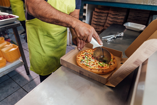Unrecognisable man running a small takeaway business in North Yorkshire, England. He is preparing food in the kitchen, cutting a pizza into slices in a takeaway box.