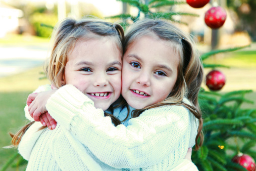 Beautiful 5 year old twins smile at the camera as they hug each other in front of a Christmas tree outside.