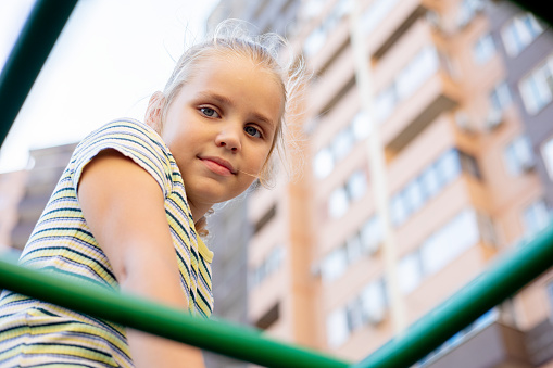 Portrait of a cute little girl. A young conqueror of the peaks is trying to overcome the steps of an alpine structure on a playground among tall multi-storey buildings.