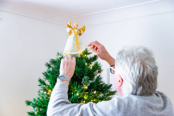 A senior man decorating his Christmas tree at home and placing the angel on top of the tree.