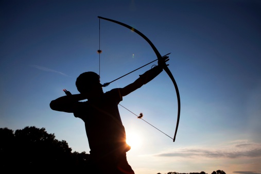 Young man in silhouette taking aim  with his bow and arrow at sunset