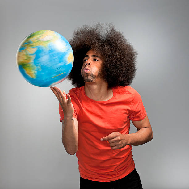 man blowing at spinning desktop globe on his fingers waist up shot of a soccer world cup fan with big afro hair in orange T-shirt blowing at a spinning globe on his fingers on gray background finger spinning globe stock pictures, royalty-free photos & images