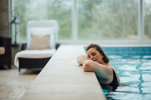 A mature woman wearing a swimming costume in an indoor home swimming pool in Ponteland, Newcastle Upon Tyne. She is at the side of the pool, relaxing with her eyes closed.
