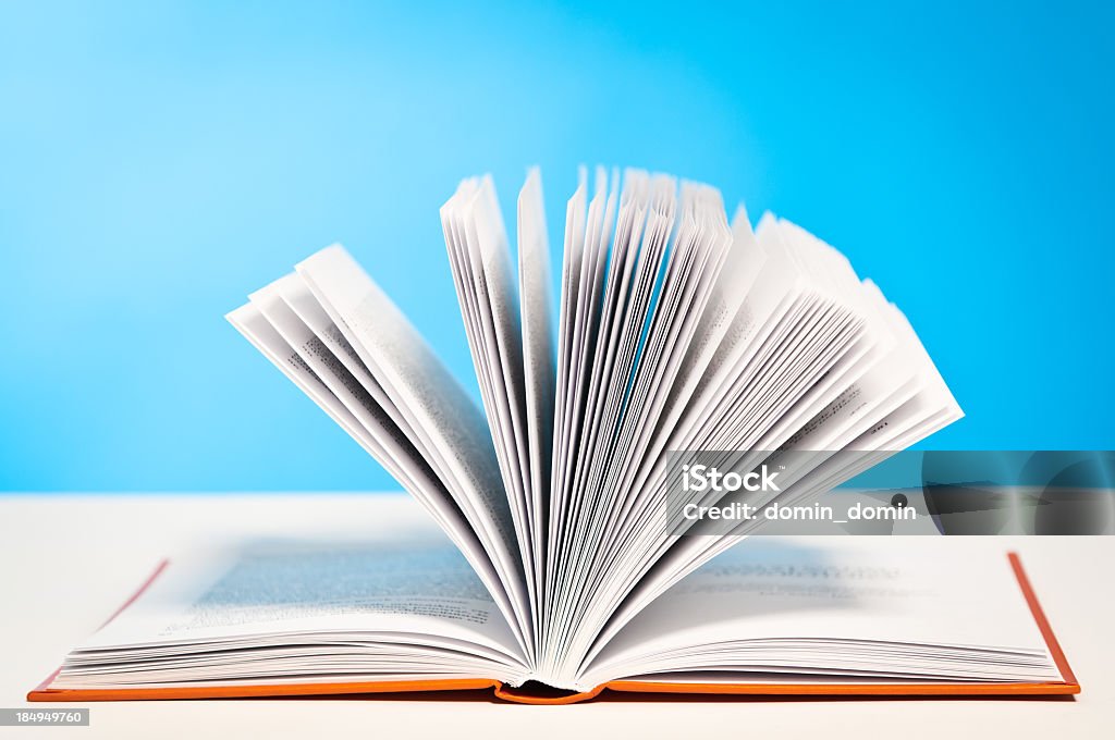 Open book, paging through pages lying on table, blue background An open book, paging through pages, lying on the white table, blue background. Copy space, horizontal, studio shot. Catalog Stock Photo