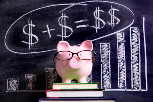 Pink piggy bank with glasses standing on books next to a blackboard with simple money math.  Sharp focus on the piggy bank with blackboard slightly blurred.  Alternative version shown below: