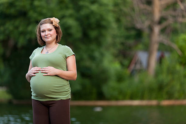 Young woman seven months pregnant with hands on her enlarged belly looks at the camera in causal clothing while standing near a park pond.