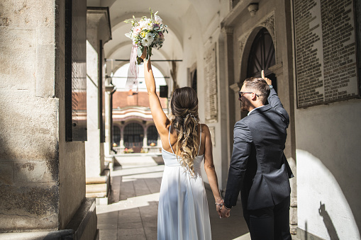 Happy couple, bride and groom holding hands and standing in front of the church before wedding ceremony, she holding wedding bouquet, back view