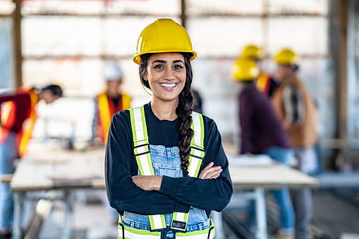 A mixed race female construction worker pauses from her work to pose for a portrait.  She is wearing proper safety gear and smiling as her coworkers meet in the background.
