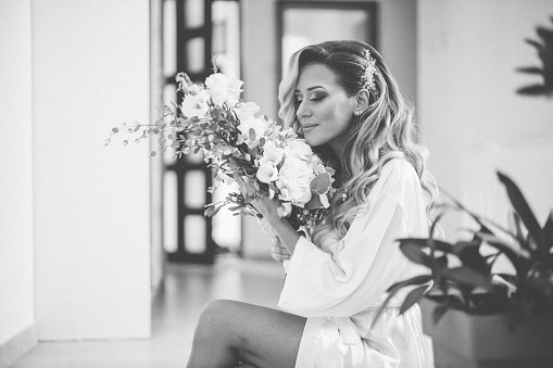Beautiful cross legged woman wearing bathrobe and sitting while holding and smelling bridal bouquet on her wedding day