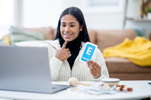 A kindergarten teacher sits in the comfort of her own home with a laptop open in front of her as she teaches virtually.  She has various musical instruments out on the table and is holding up a cue card as she teaches the letters and sounds of the alphabet.