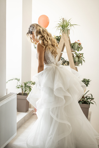 Young beautiful elegant bride holding bridal bouquet and posing in living room in front of ladder with potted plants, side view