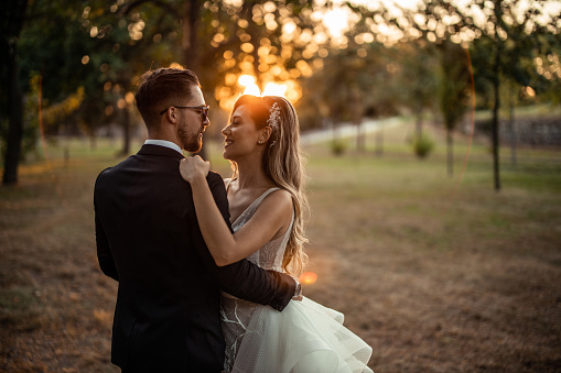 Beautiful smiling bride and groom gently embracing in nature on grass area during sunset