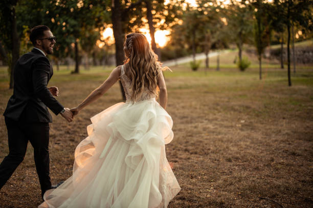 Laughing married couple holding hands and walking through grass field in park during sunset, back view