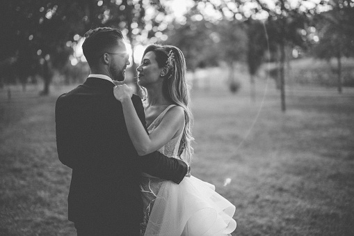 Beautiful smiling bride and groom gently embracing in nature on grass area during sunset, black and white photo