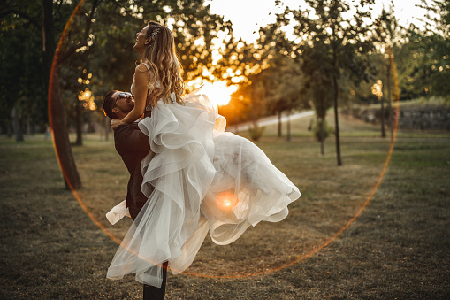 Beautiful bride laughing while groom lifting her on grass field during sunset
