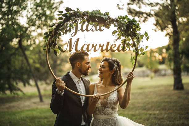 Young married couple, bride and groom holding floral circle with just married letters inside and looking each other on grass field