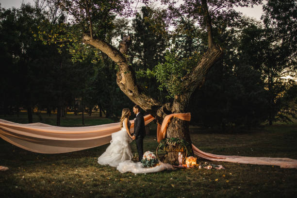 Smiling bride and groom standing and holding hands while looking each other on grass field under tree in decorative ambient with candles and flowers
