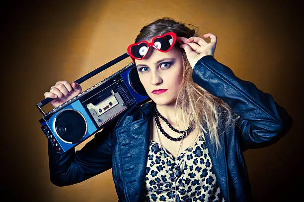 A beautiful teenage girl dressed in 80's fashion listening to a cassette boom box and lifting her heart shaped glasses.