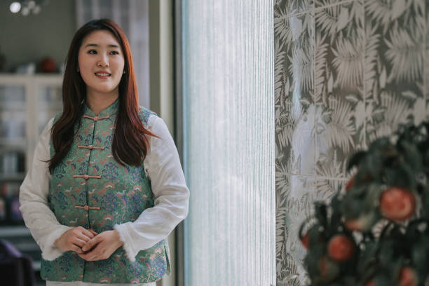 donna del capodanno cinese con cheongsam tradizionale che distoglie lo sguardo sorridente in soggiorno - cheongsam chinese culture indoors looking at camera foto e immagini stock