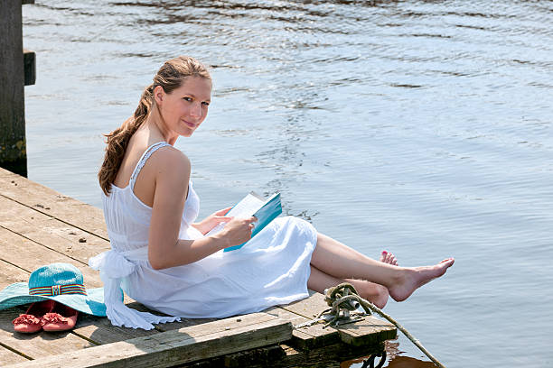 Resubmission of file #16562922 as approved by HQ. email CIA@istockphoto.com for detailsside view on a woman with barefeet in a white sundress reading a magazine sitting on wooden pier at the waterfront looking at the camera