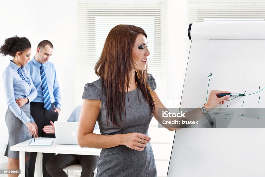 Business training Focus on the young businesswoman drawing graph on the flip chart with her colleagues working in the background. Adult Stock Photo