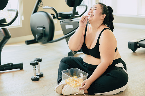 Binge Eating Disorder (BED), happy fat women laugh enjoy unstopable eating chips snack food during relax brake from exercise in fitness sport club.