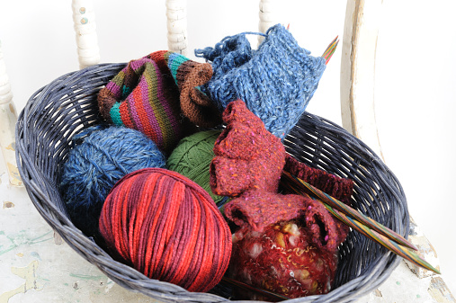 balls of yarn, multi-colored knitting needles and handmade sock and glove in a basket sitting on a white chair against a white background