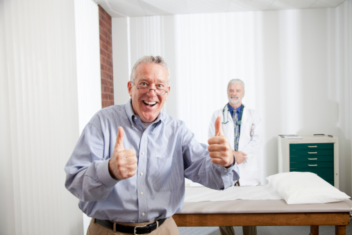 A patient in his late 50's early 60's gives a thumbs up with a male doctor in the background.