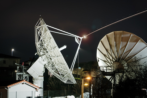 Two satellite ground station antennas at night. Line of light coming from the antenna, originally from an airplane, digitally modified
