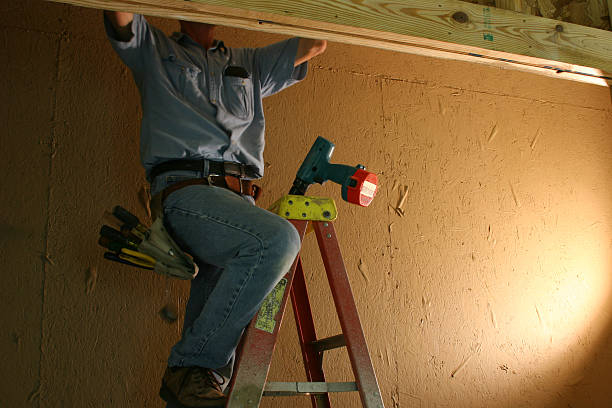 Caucasian Electrician working in the closet with a clamp on light to help him see.  He is hanging a new light fixture in a newly constructed closet.  Focus is on his portable drill that is in the top of the ladder.  