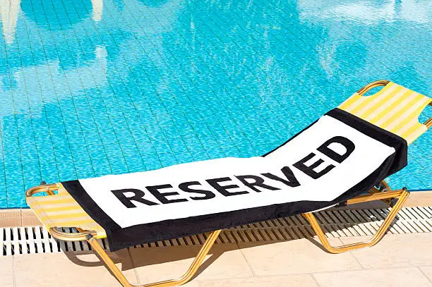 A beach towel with the word 'Reserved' spread onto a sun lounger beside a swimming pool