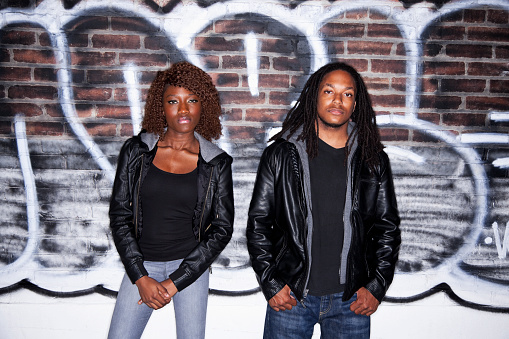 Young black couple (20s) standing in front of brick wall covered with grafitti.