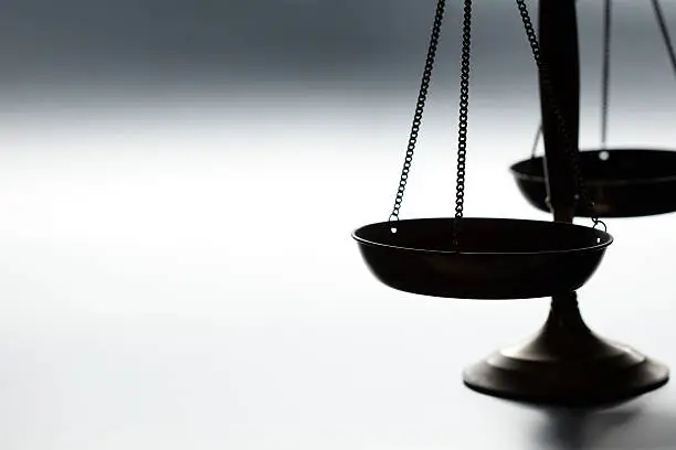 A justice scale on a simple gray background.  The scale is partially silhouetted as a strong backlight obscures some of the finer details.  Scale is place on far right hand side of image leaving ample negative space for copy. Image can be flipped horizontal to accommodate alternative composition needs.