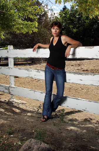 A clear Photograph of a brown haired, Mature Woman by a fence, wearing blue denim jeans, black tank top, brown belt, black sandals, a pony tail, and red nail polish. She is leaning on a fence. She doesn't look very happy but doesn't look mad either.