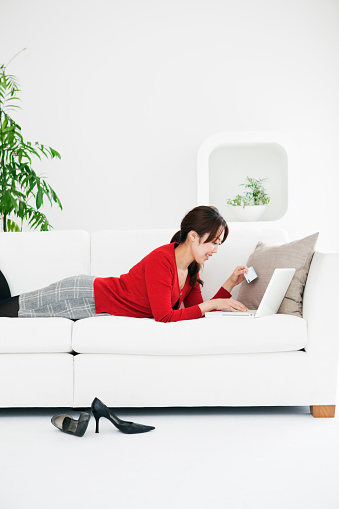 Subject: A young Asian woman laying on couch shopping on line using her credit card and laptop computer in the living room of her home.