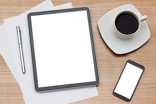 Tablet, smartphone, stylus and cup of coffee on a white saucer on a wooden table.  The tablet has a black border with a white screen and is placed on top of white pieces of paper with a silver stylus pen placed to the left of the stylus.  A black bordered smartphone with a shiny, reflective screen is placed toward the bottom right of the image.  A white cup of coffee is placed above the smartphone and is sitting on a square white saucer.  The electronics are placed on a wooden table.